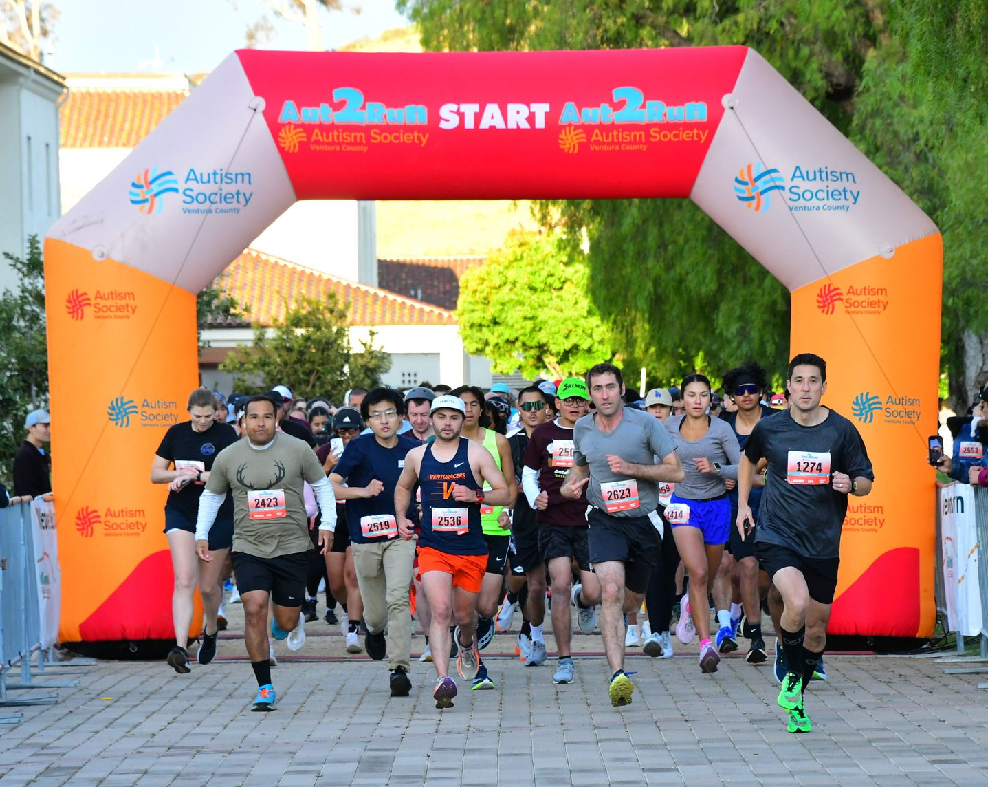 Runners starting a charity race beneath a red-and-orange start arch on a sunny outdoor path