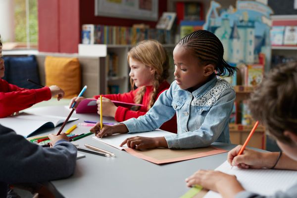Bambini che scrivono con matite colorate a un tavolo in classe, con un quaderno aperto e dei libri nelle vicinanze.