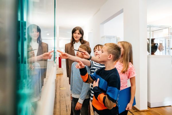 Dei bambini indicano una teca di vetro in un museo, mentre una donna si trova nelle vicinanze, in una galleria luminosa.