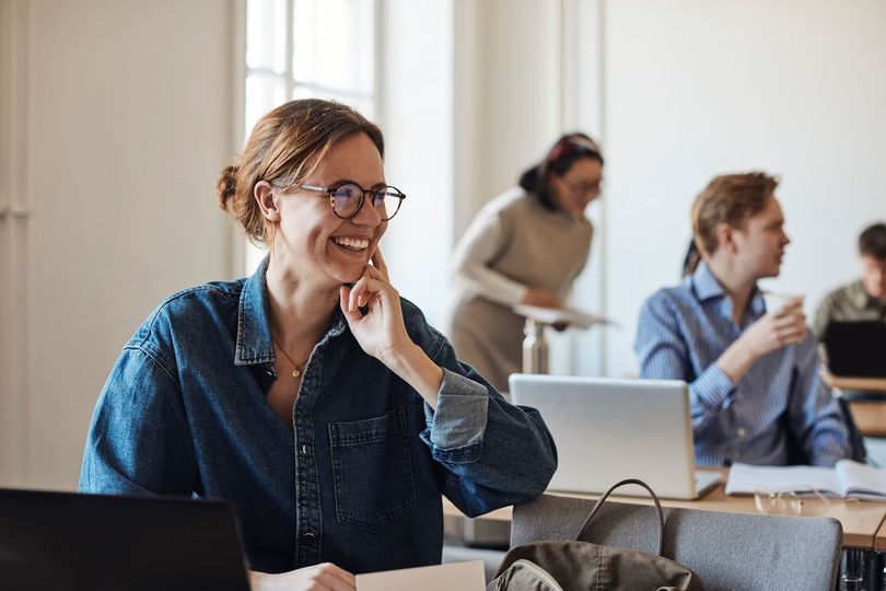 Persona sorridente con gli occhiali seduta a una scrivania in ufficio, con i colleghi sfocati sullo sfondo.