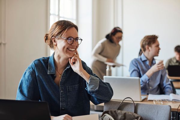 Persona sorridente con gli occhiali davanti a un computer portatile in un ufficio luminoso, con i colleghi sullo sfondo.