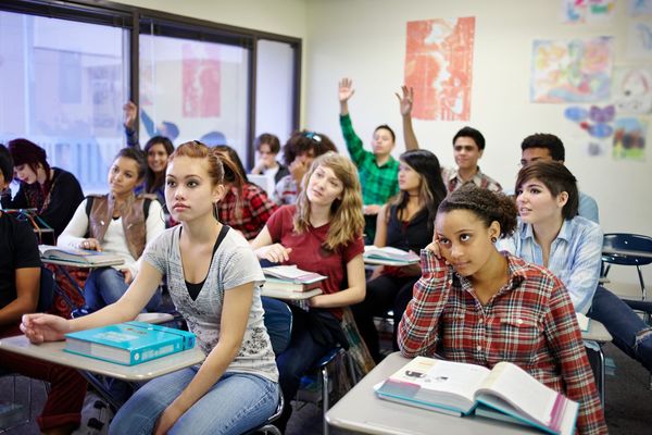 Studenti seduti in un'aula, che alzano la mano durante una lezione con libri aperti sui banchi.