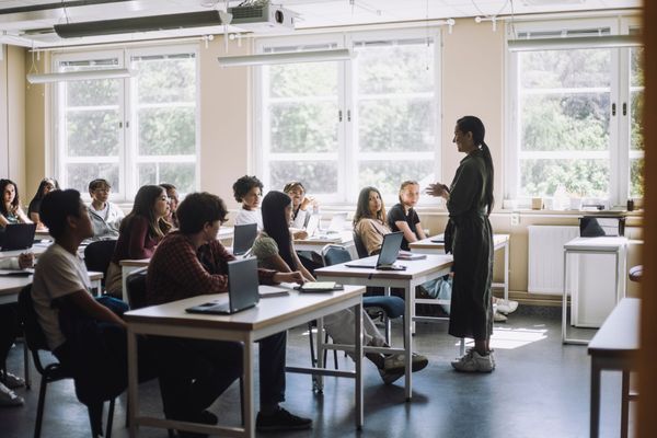 Un'insegnante si rivolge agli studenti in un'aula luminosa con grandi finestre e banchi.
