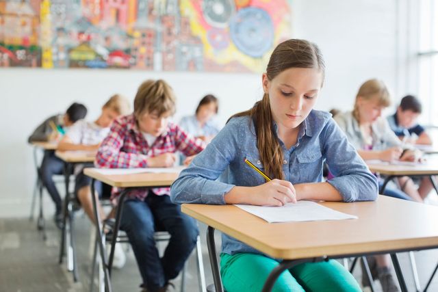 Studenti che sostengono un esame in un'aula, con una ragazza in camicetta blu che scrive a una scrivania di fronte a loro.