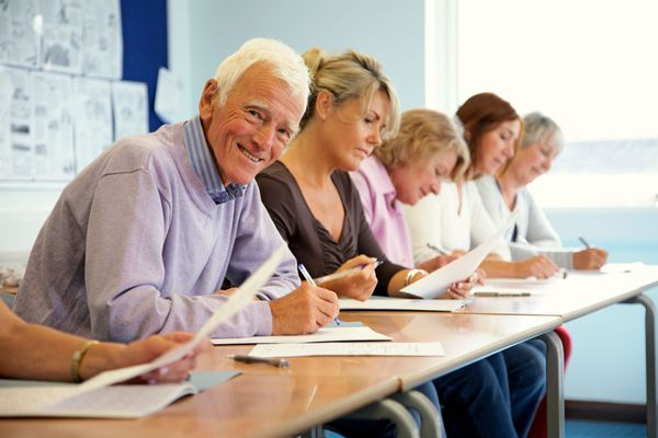 Gruppo di persone sedute attorno a un lungo tavolo, intente a scrivere su dei fogli di carta in un'aula o sala riunioni luminosa.