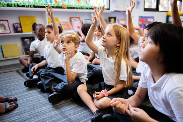Bambini seduti sul tappeto di un'aula con le mani alzate durante una lezione.