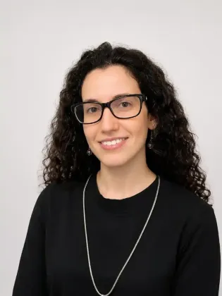 Ritratto di una persona sorridente con capelli scuri e ricci, occhiali e una maglietta nera su sfondo chiaro.