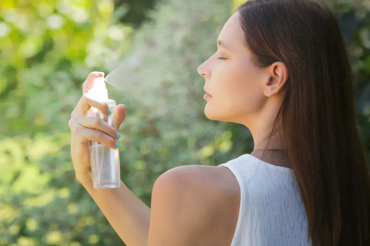 Mujer aplicándose protector solar al aire libre bajo la luz del sol, sosteniendo un pulverizador cerca del hombro.