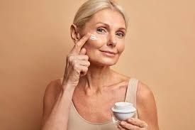 Persona aplicándose crema en la mejilla mientras sostiene un pequeño frasco sobre un fondo beige.