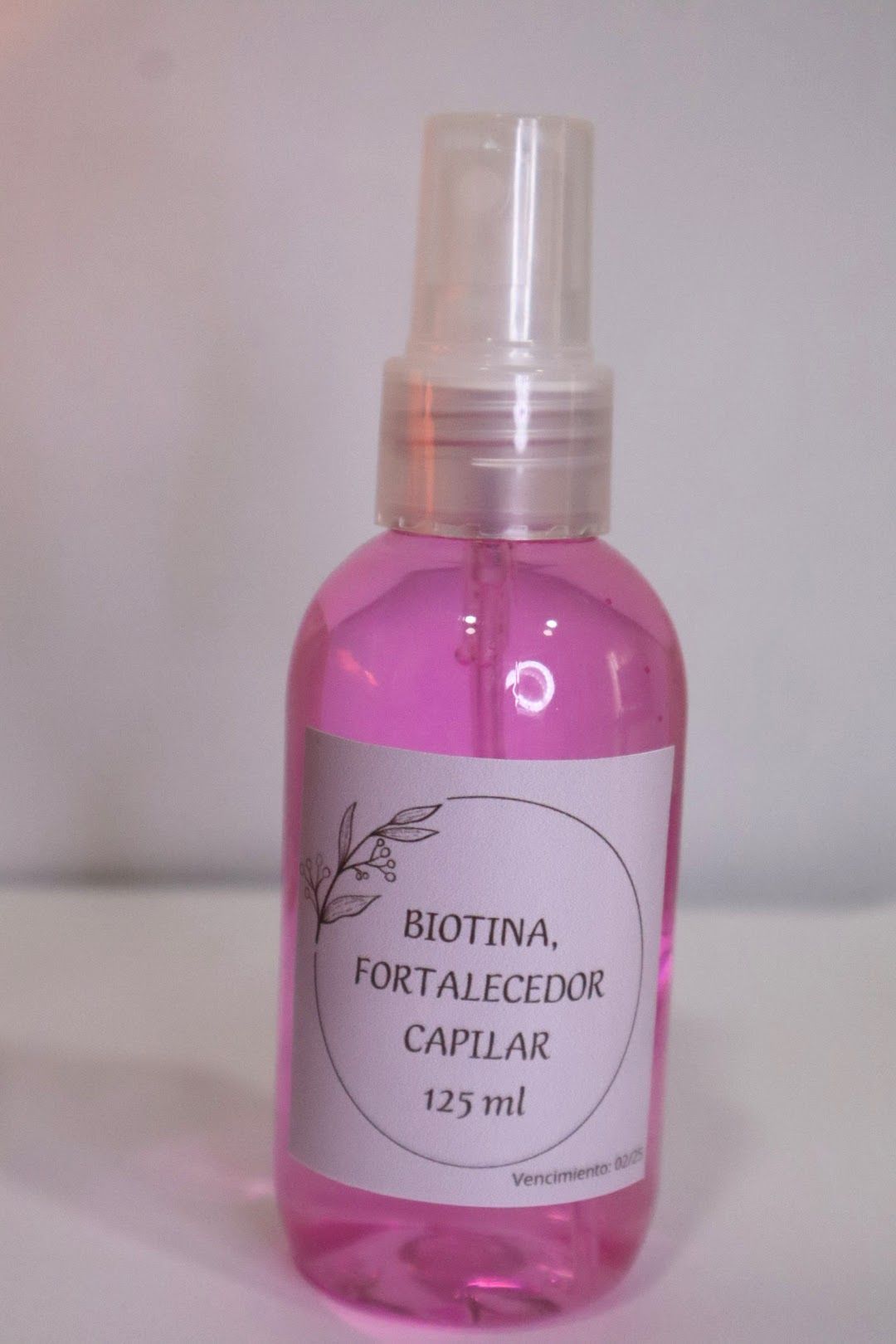 Frasco de spray para el cuidado del cabello de color rosa con la etiqueta “Biotina, Fortalecedor Capilar” sobre una superficie blanca.