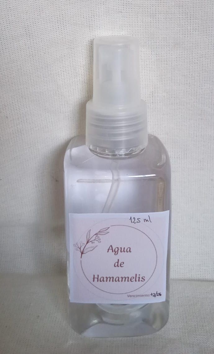 Botella de spray transparente con la etiqueta “Agua de Hamamelis” sobre un fondo blanco texturizado.