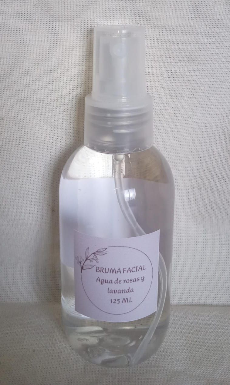 Botella pulverizadora transparente con una etiqueta de color lavanda sobre un fondo de tela blanca texturizada.