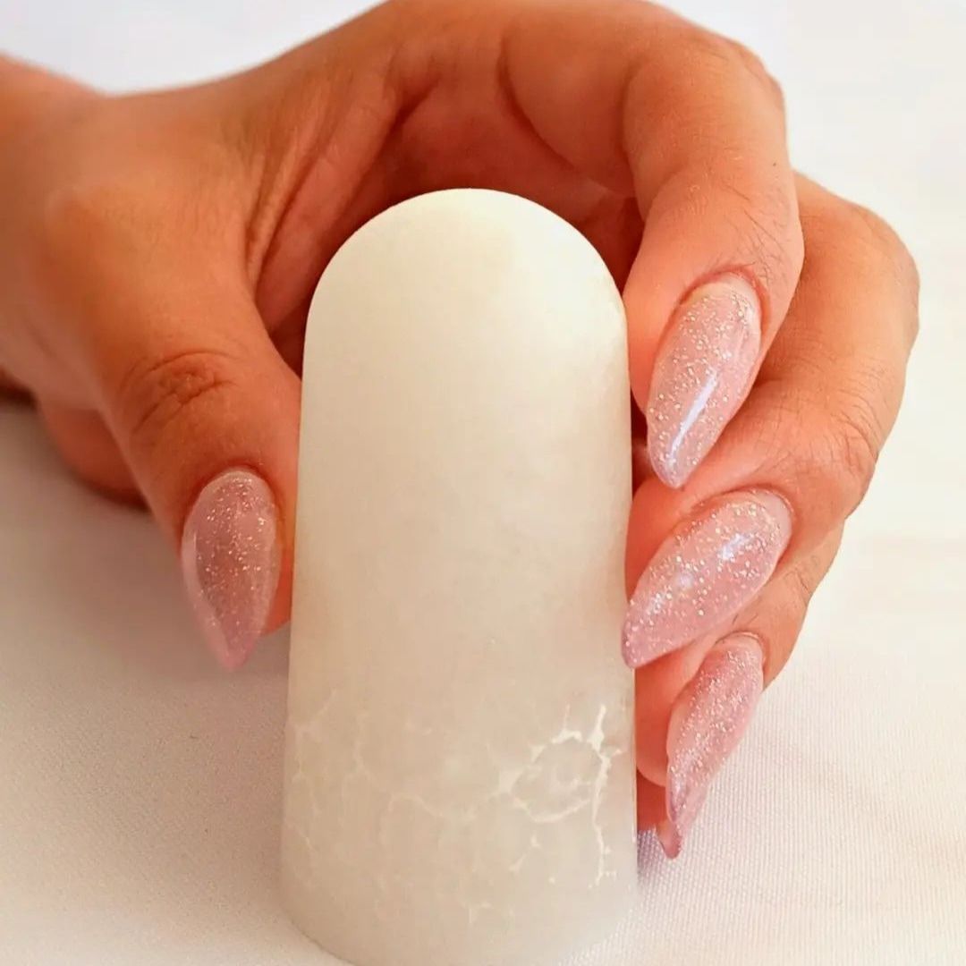 Mano sosteniendo un soporte blanco abovedado para exhibir uñas con manicura de purpurina color nude.