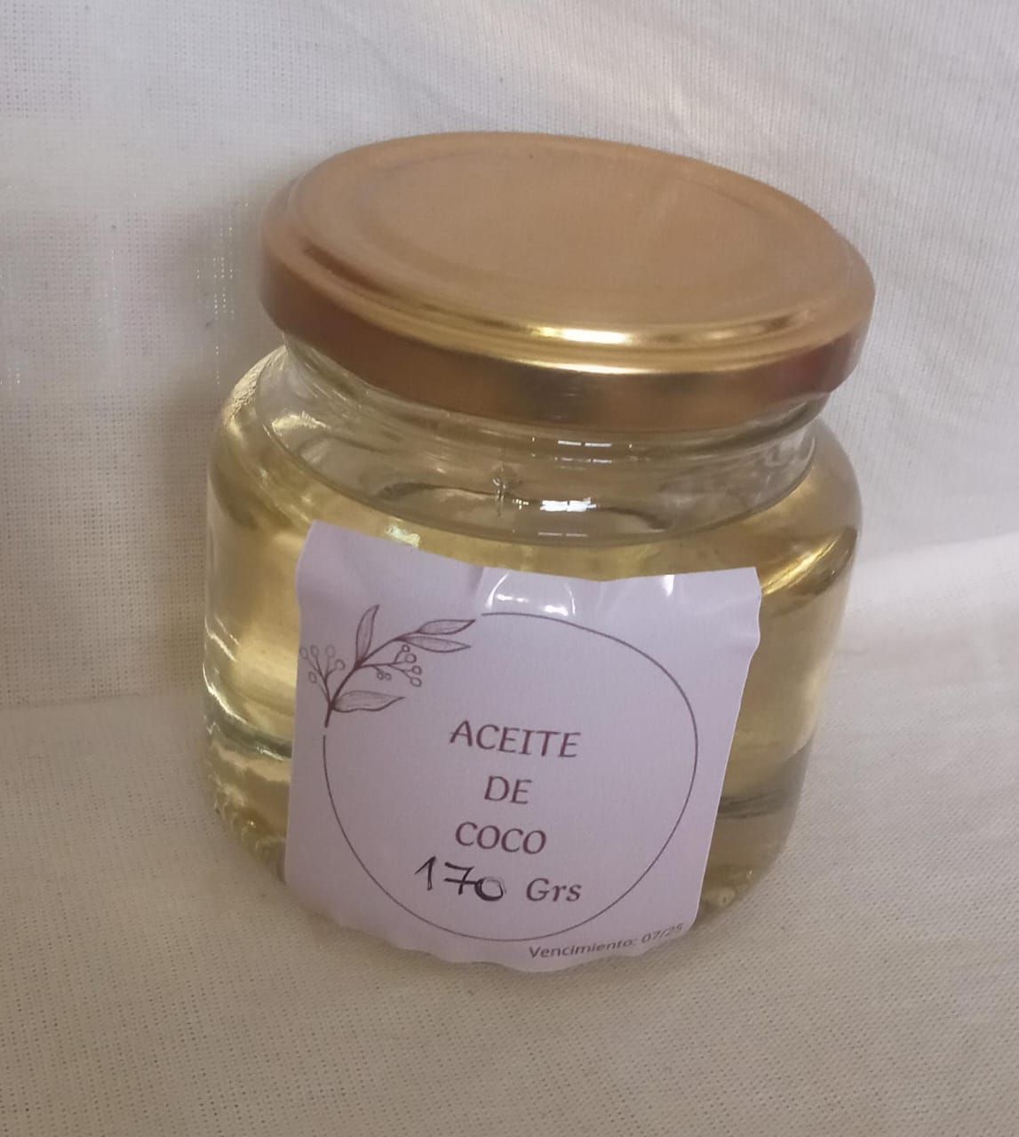 Frasco pequeño de vidrio con tapa dorada etiquetado “Aceite de coco 170 Grs” sobre una superficie de tela clara.