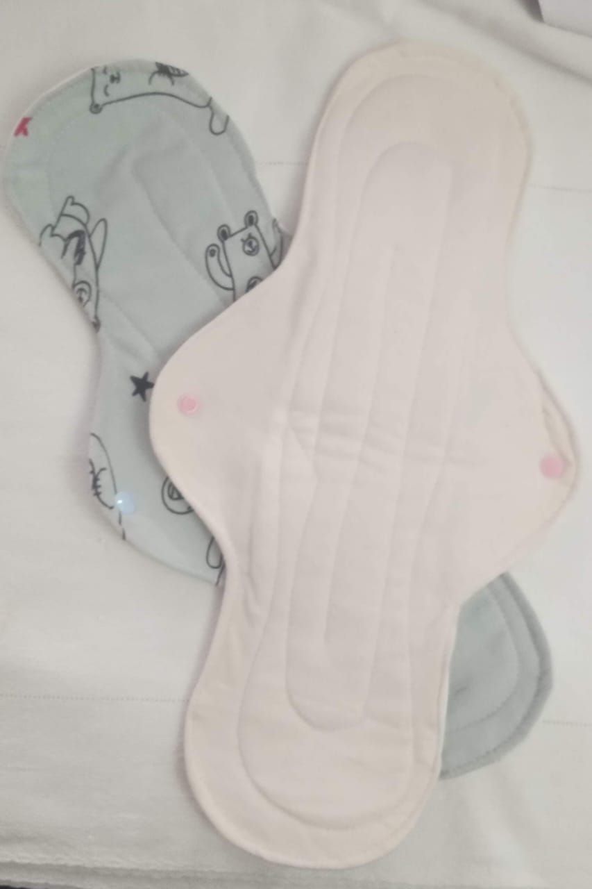 Dos compresas de tela reutilizables, una azul claro con pequeños estampados y otra blanca, superpuestas sobre una superficie blanca.