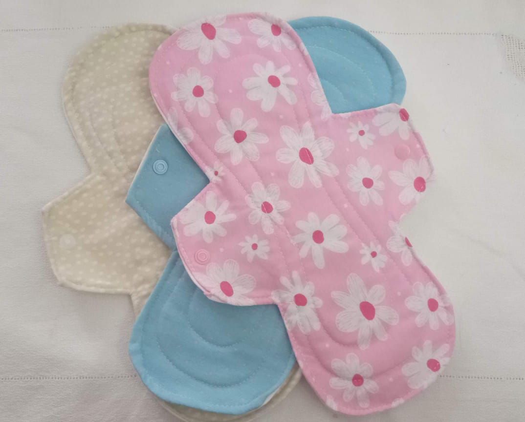 Compresas de tela reutilizables apiladas en rosa floral, azul y crema sobre un fondo blanco.
