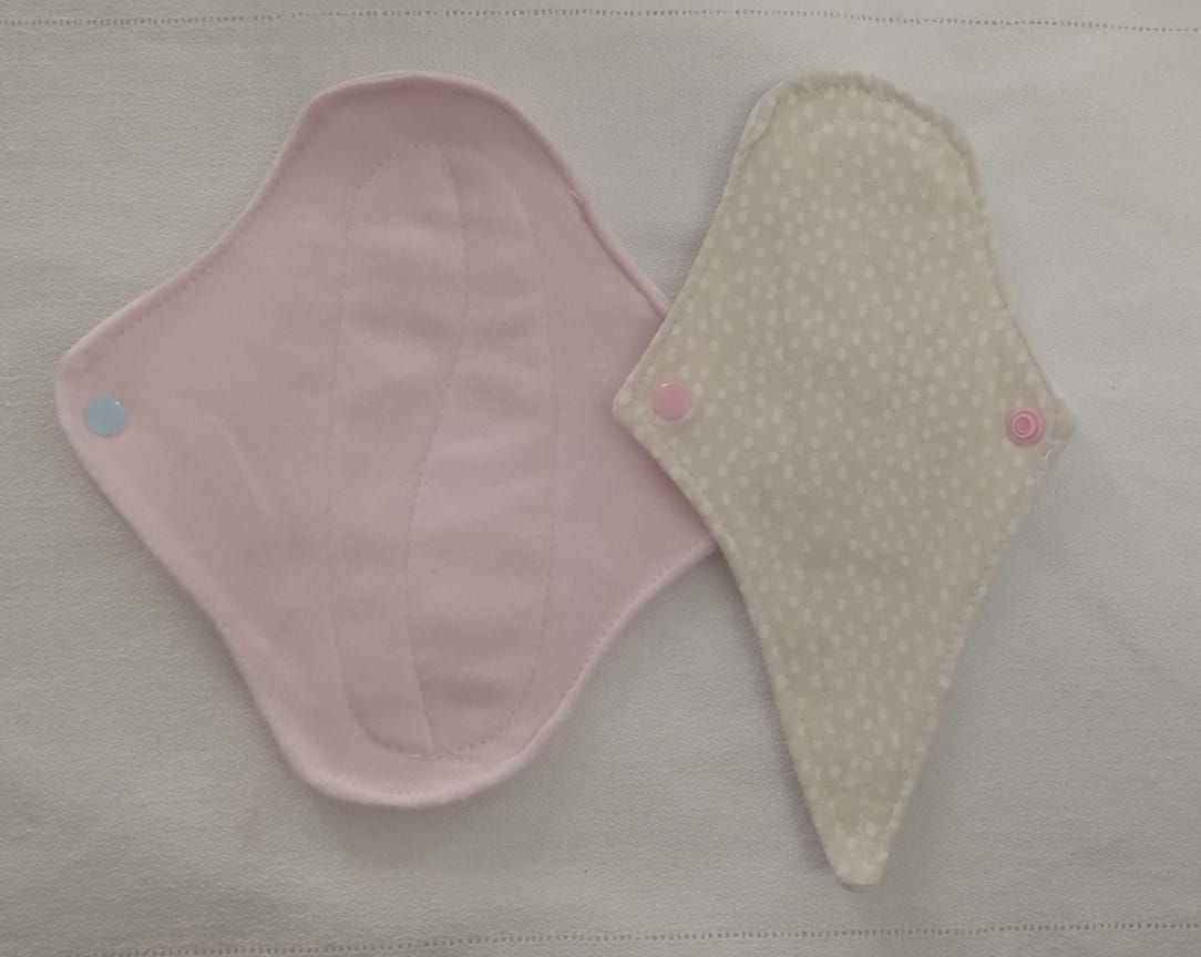 Dos compresas de tela reutilizables, una rosa y otra color crema con puntos rosas, colocadas una al lado de la otra sobre una superficie clara.
