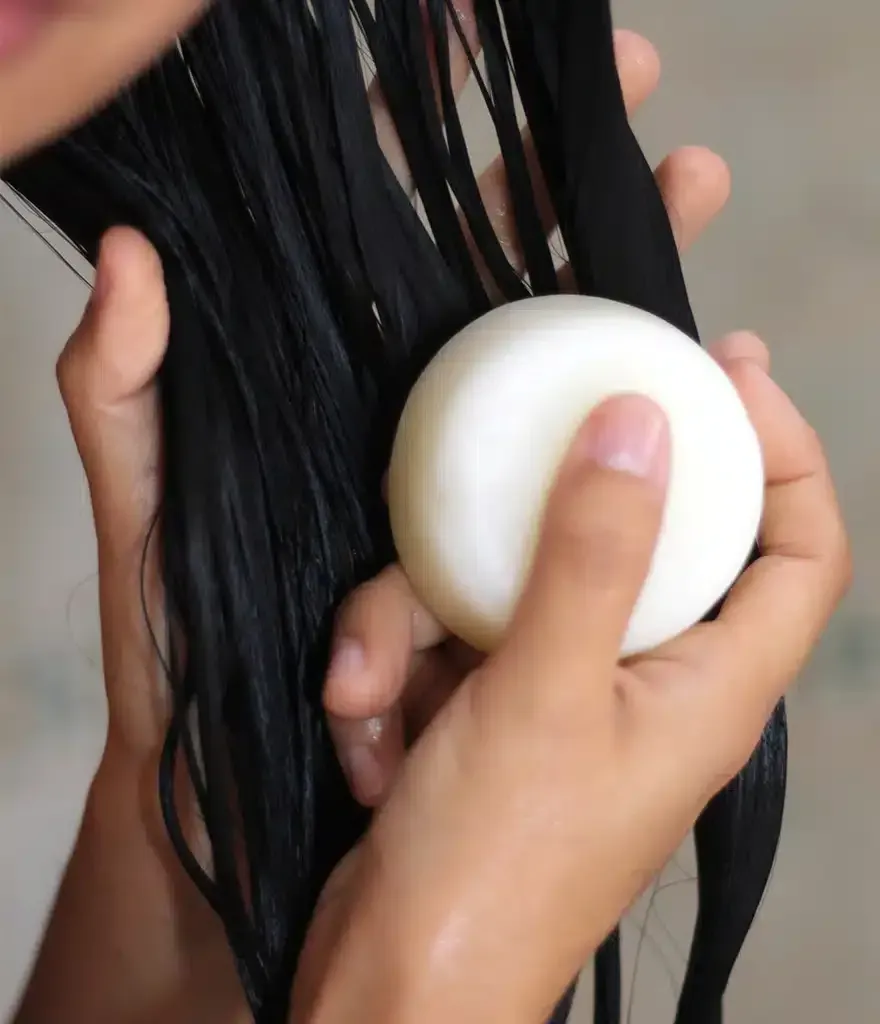 Manos lavándose el pelo largo y negro con una pastilla de champú blanca.