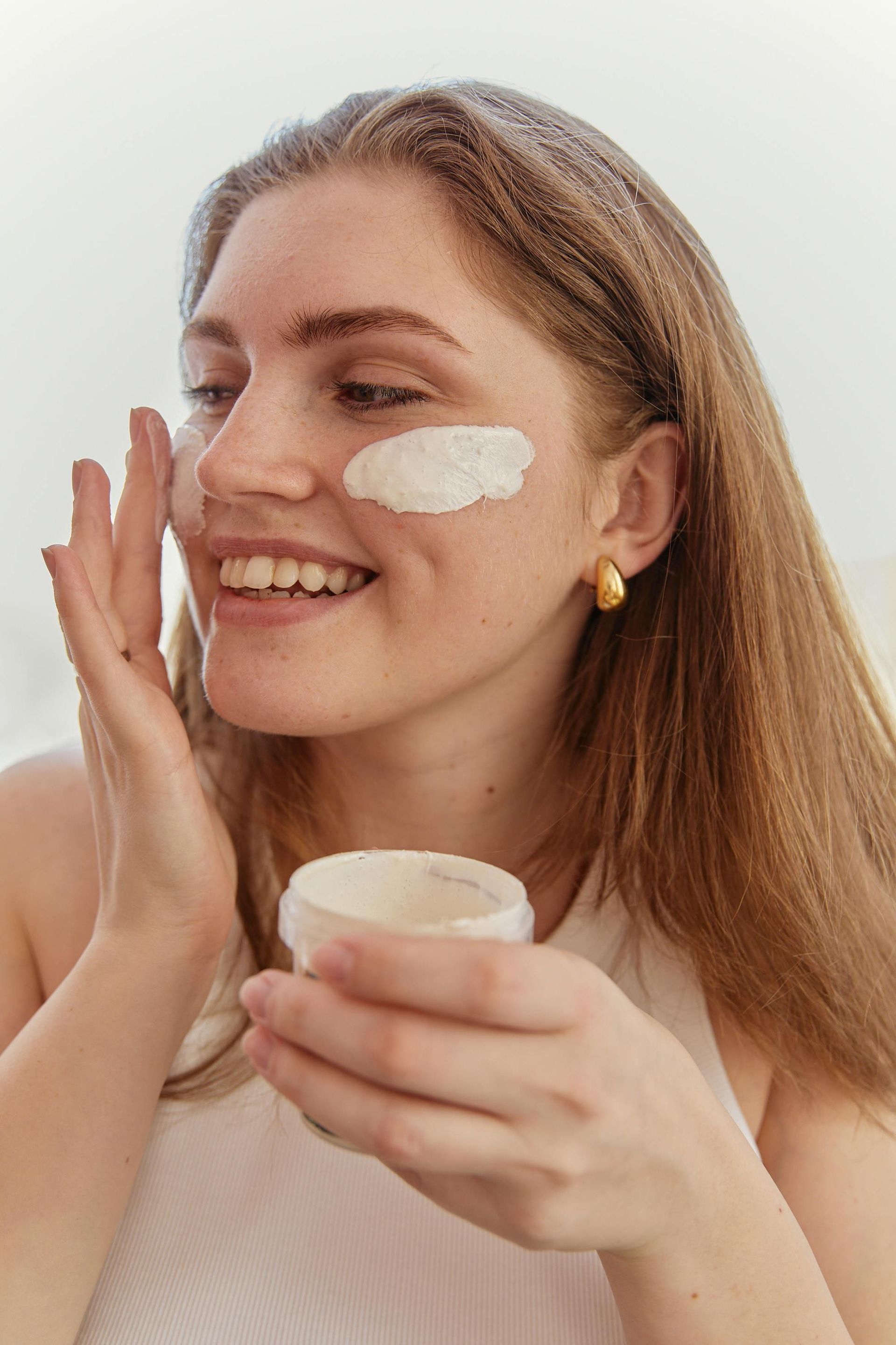 Mujer aplicándose crema en la mejilla mientras sostiene un frasco de crema para el cuidado de la piel.