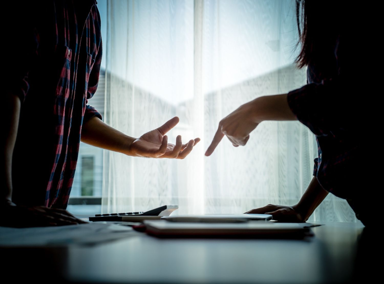 Two people at a table, arguing. One points at a tablet, the other gestures with open hand. Sunlight streams through a window.