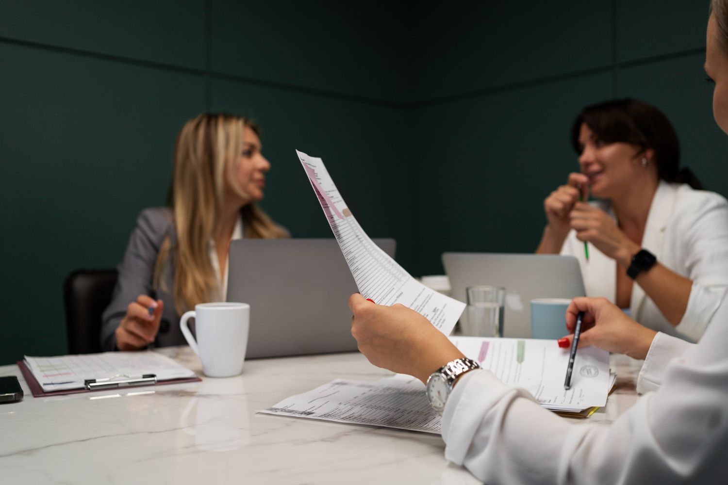 Three people in a meeting room, reviewing papers and using laptops. One person holds the papers and others converse.