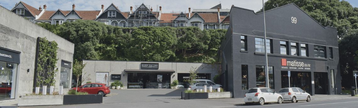 Exterior view of modern buildings with cars parked in front and houses on a hill in the background.