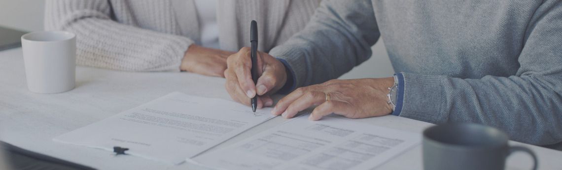 Person signing documents at a table; a pen in hand, next to a mug and other person.
