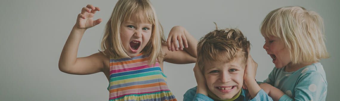 Three children, two girls and a boy, yelling and covering their ears, against a light gray background.