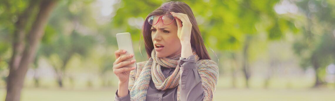 Woman looking at a smartphone with a concerned expression, touching her head outdoors.