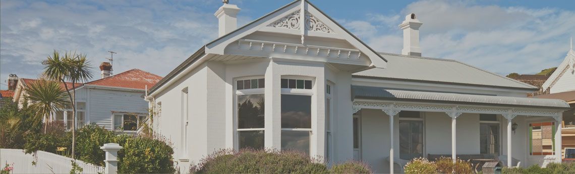 White house with decorative trim and porch. Other houses and greenery in the background.
