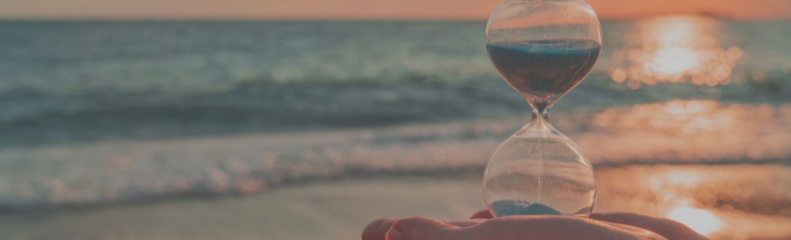 Hourglass held in front of ocean at sunset, sand flowing from top to bottom.
