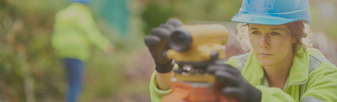 A person in safety gear using a surveying instrument in an outdoor setting.
