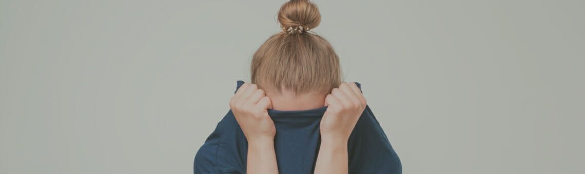 Person wearing a dark blue shirt, pulling it over their head. Topknot hairstyle visible. Plain backdrop.