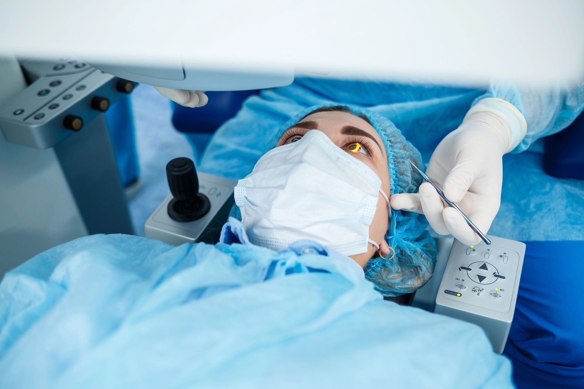 Un paziente, con indosso camice, mascherina e cuffia chirurgica, giace sotto un apparecchio laser per la chirurgia oculare, mentre un chirurgo con i guanti si prepara per un intervento.