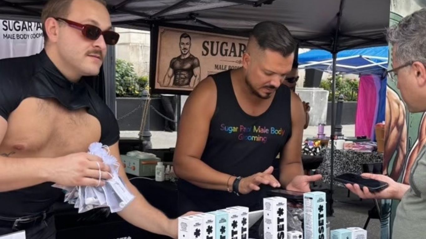 Three people at an outdoor market booth, examining items under a tent with a “SUGAR” sign.