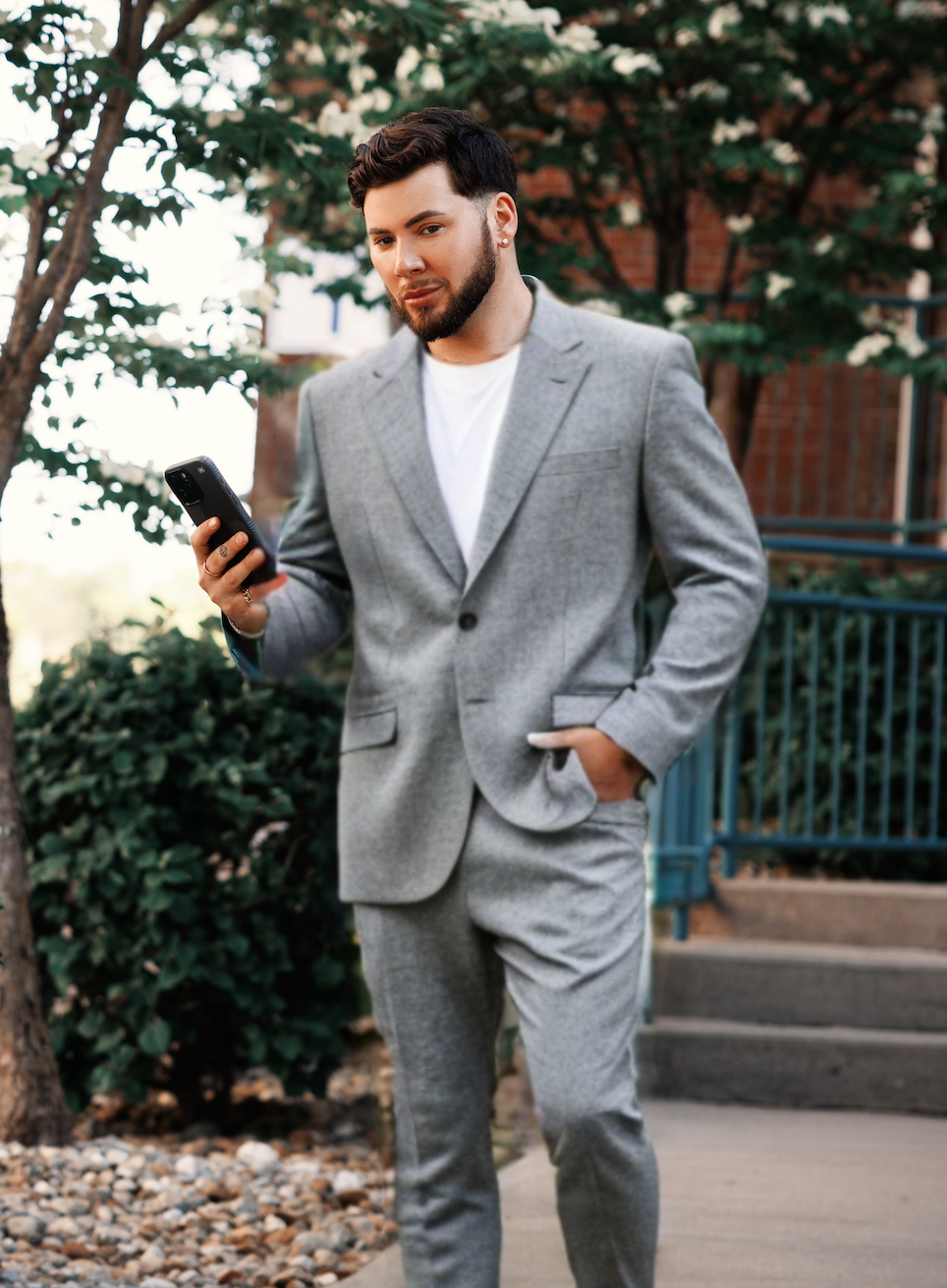 A person in a light grey suit and white t-shirt walks outdoors while looking at a smartphone held in their hand.