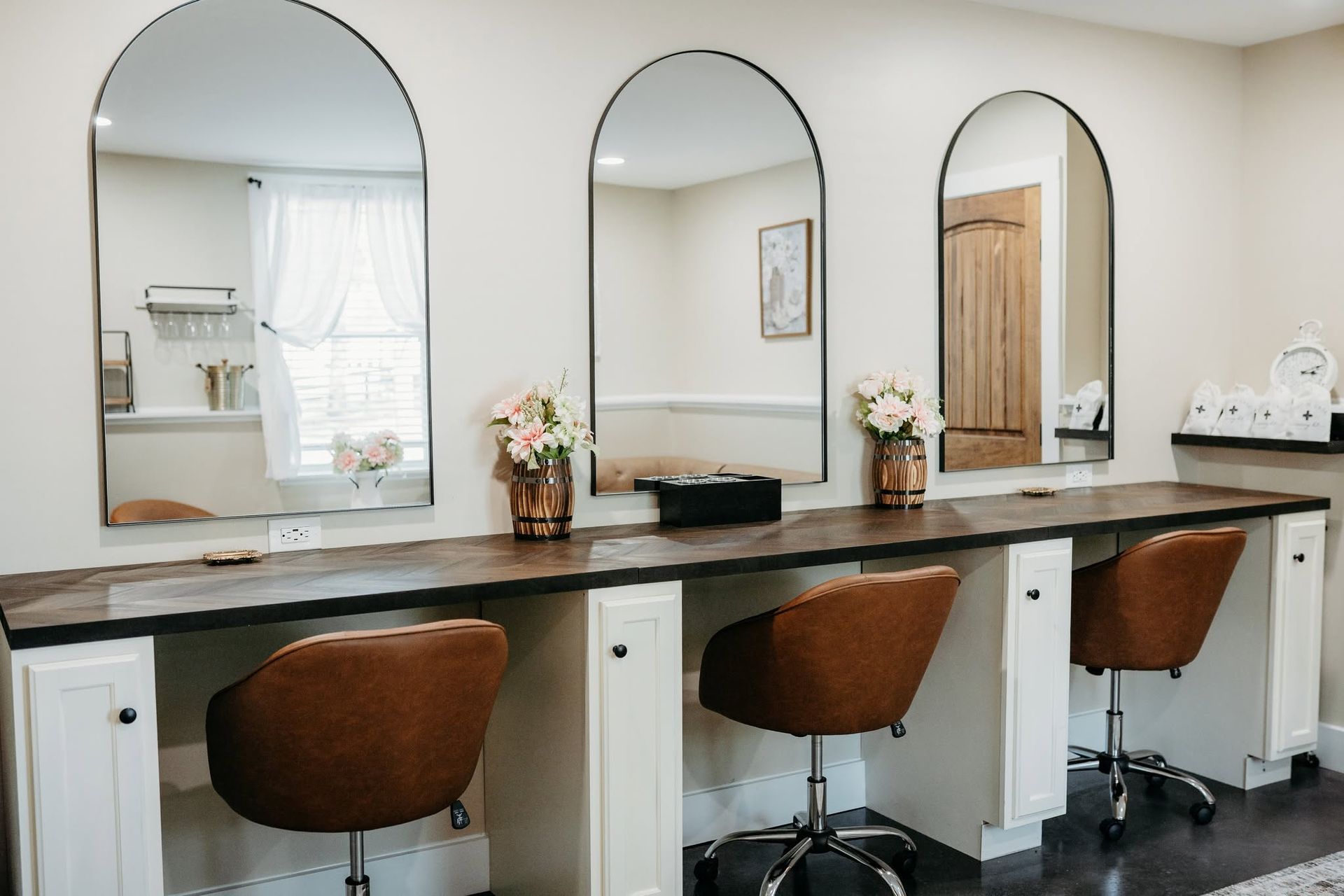 Salon vanity with three mirrors, brown stools, and flowers on a white countertop