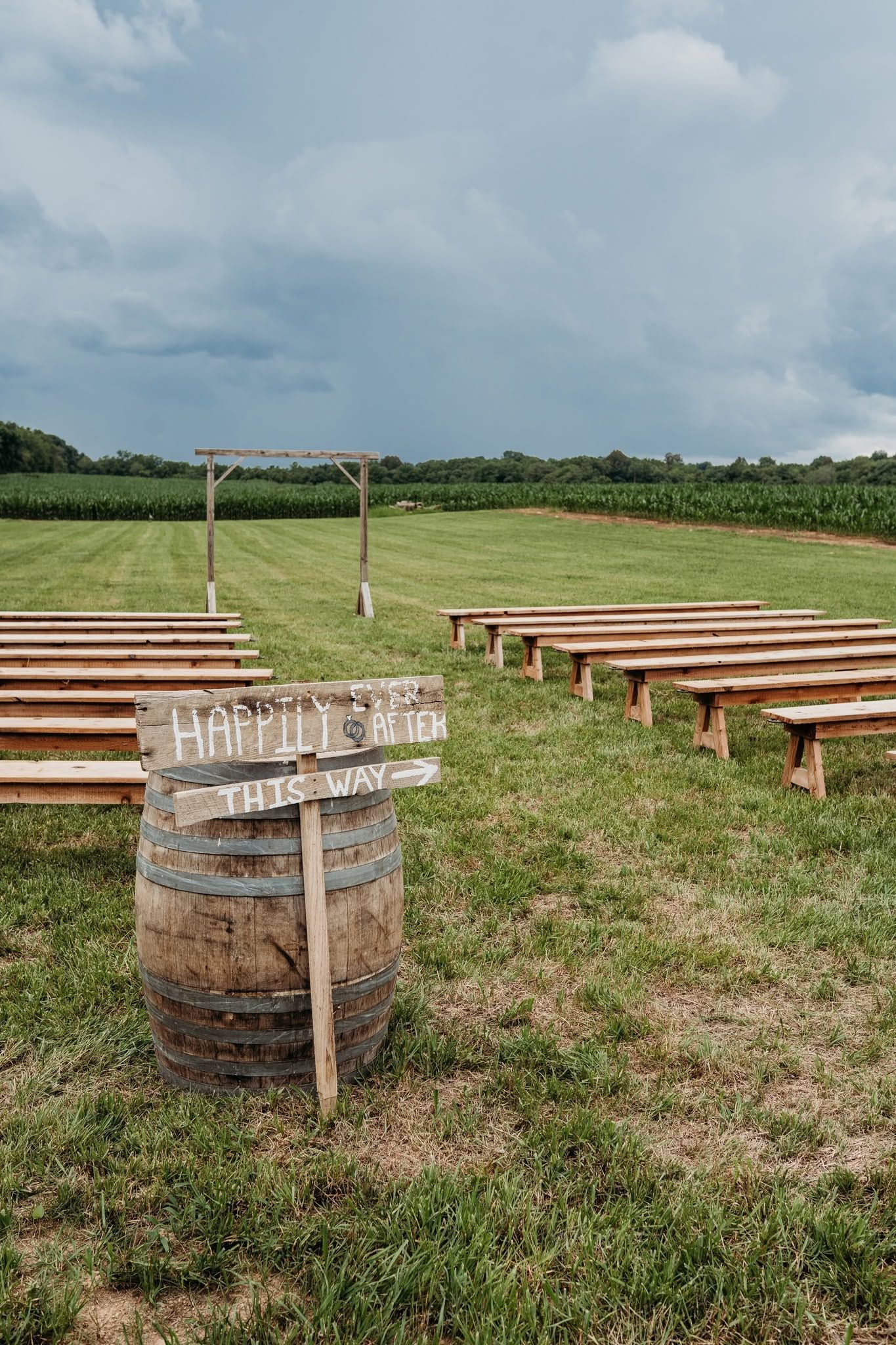 Outdoor picnic setup with wooden benches and a rustic sign in a grassy field under cloudy sky