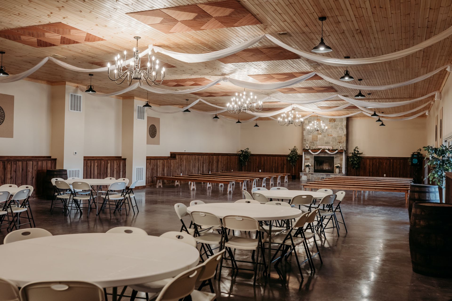 Empty banquet hall with round tables, folding chairs, chandeliers, and draped ceiling decor