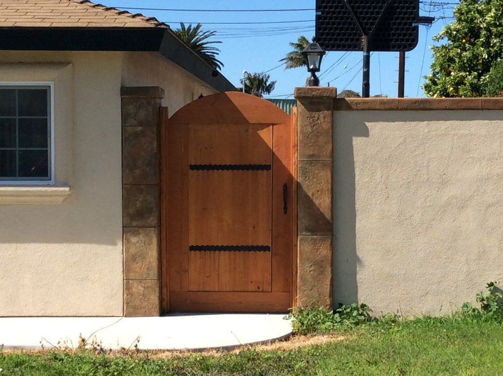 Wooden arched gate in a stucco wall, flanked by stone columns and a beige wall; sunny day.