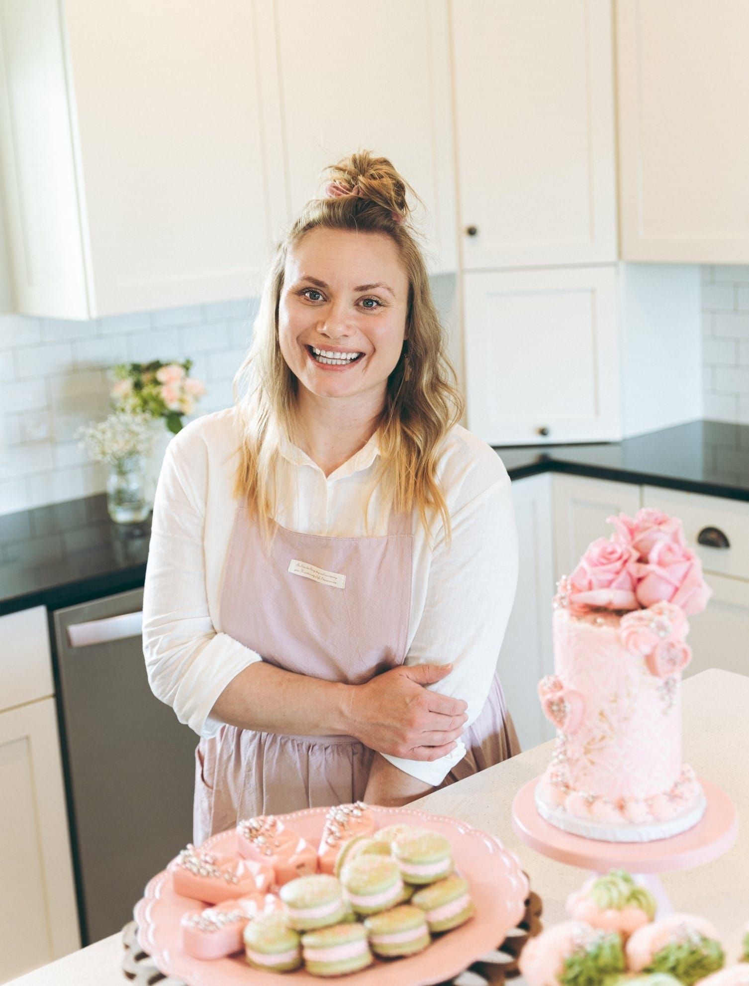 Woman in pink apron smiles near a decorated pink cake and pastries on a kitchen counter.