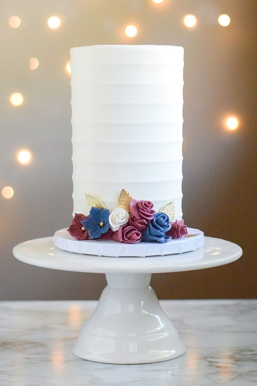White cake with textured frosting, decorated with blue, burgundy, and gold flowers on a white cake stand.