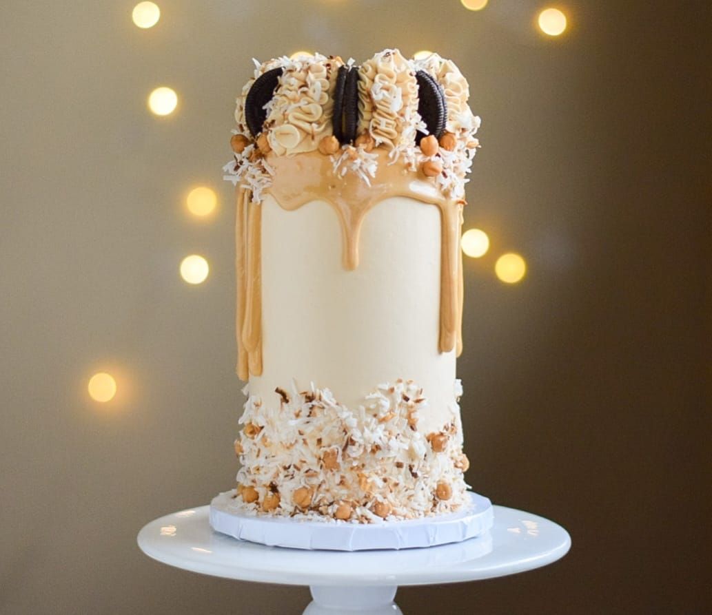 Tall, cylindrical cake with tan frosting, caramel drip, and cookie and coconut topping on a white cake stand.