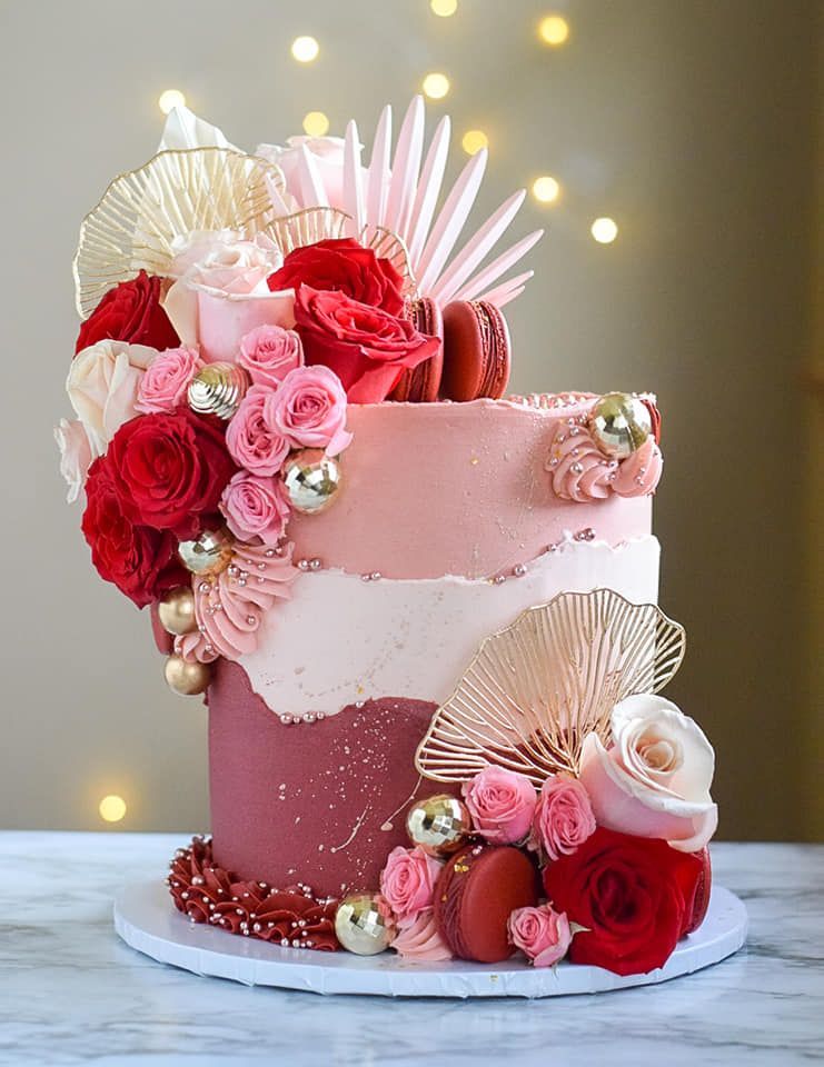 Two-tiered cake with red and pink roses, gold accents, and macarons on a white base.