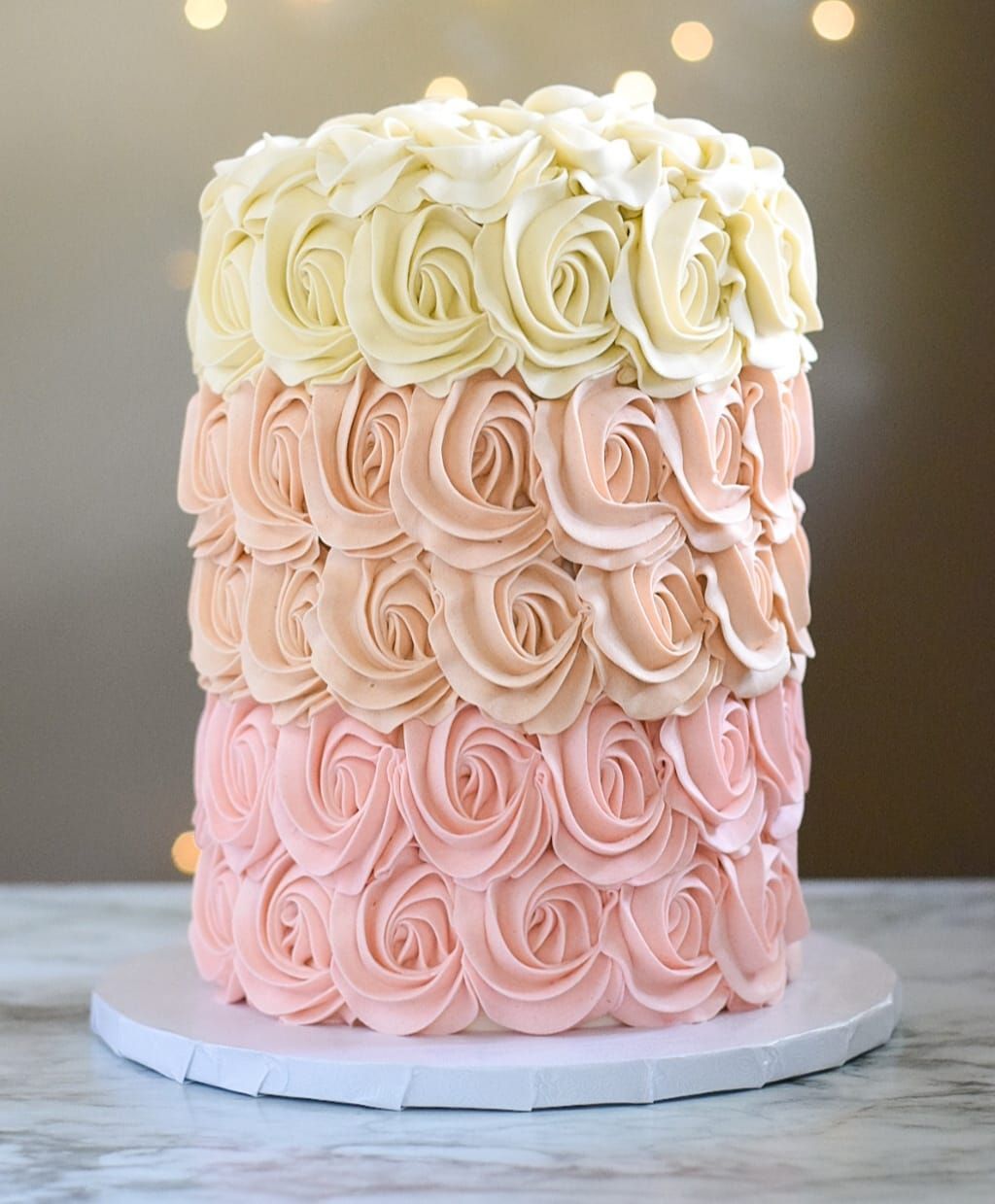 Three-tiered cake with frosting roses, ranging from pink to cream, on a white cake board.