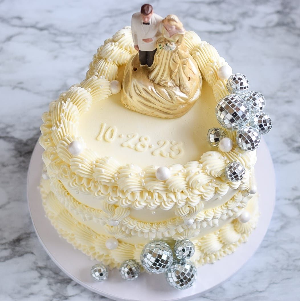 Heart-shaped wedding cake with cream frosting, bride & groom topper, date, & disco ball decorations.
