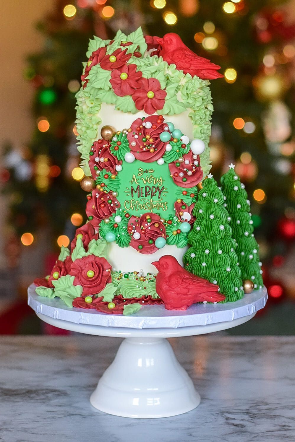 Christmas cake on a pedestal, decorated with red birds, flowers, and greenery against a Christmas tree.