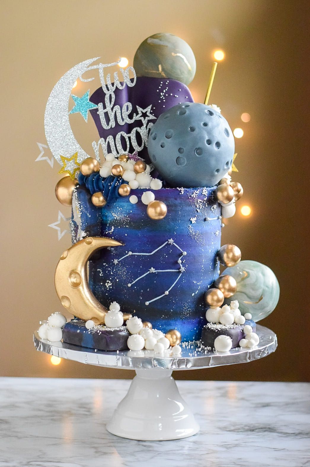 Space-themed cake with galaxy frosting, planets, gold moons, star constellations, on white cake stand.
