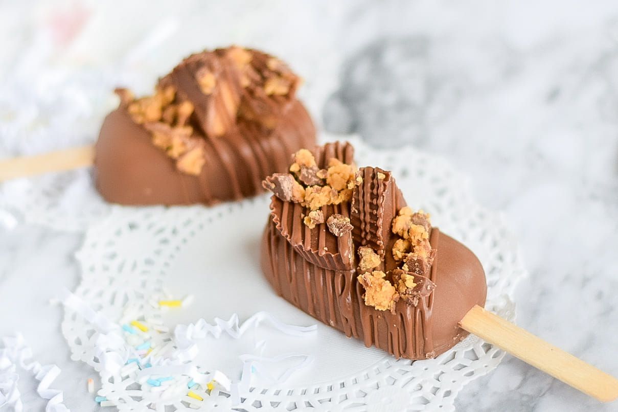 Chocolate-covered desserts on sticks, garnished with peanut butter cups, on a lace doily, against a blurred background.