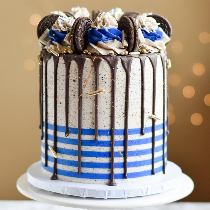 Cake with chocolate drips, blue stripes, Oreo cookies, and cream frosting on a white cake stand.